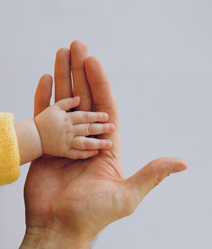 Crop anonymous baby in warm wear holding hand on palm of crop parent while standing against white background in studio